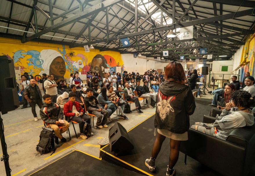 Visão em plano médio de jovens sentados em fileiras horizontais de cadeiras no galpão da Vila Itororó, assistindo a uma palestra. No primeiro plano à direita, a palestrante está de costas, em pé sobre um palco baixo, falando para o público. Ao fundo, destacam-se as paredes do interior do galpão na tonalidade amarela com murais de grafite, enquanto outras pessoas circulam livremente pelo evento sob um teto de estrutura metálica exposta.